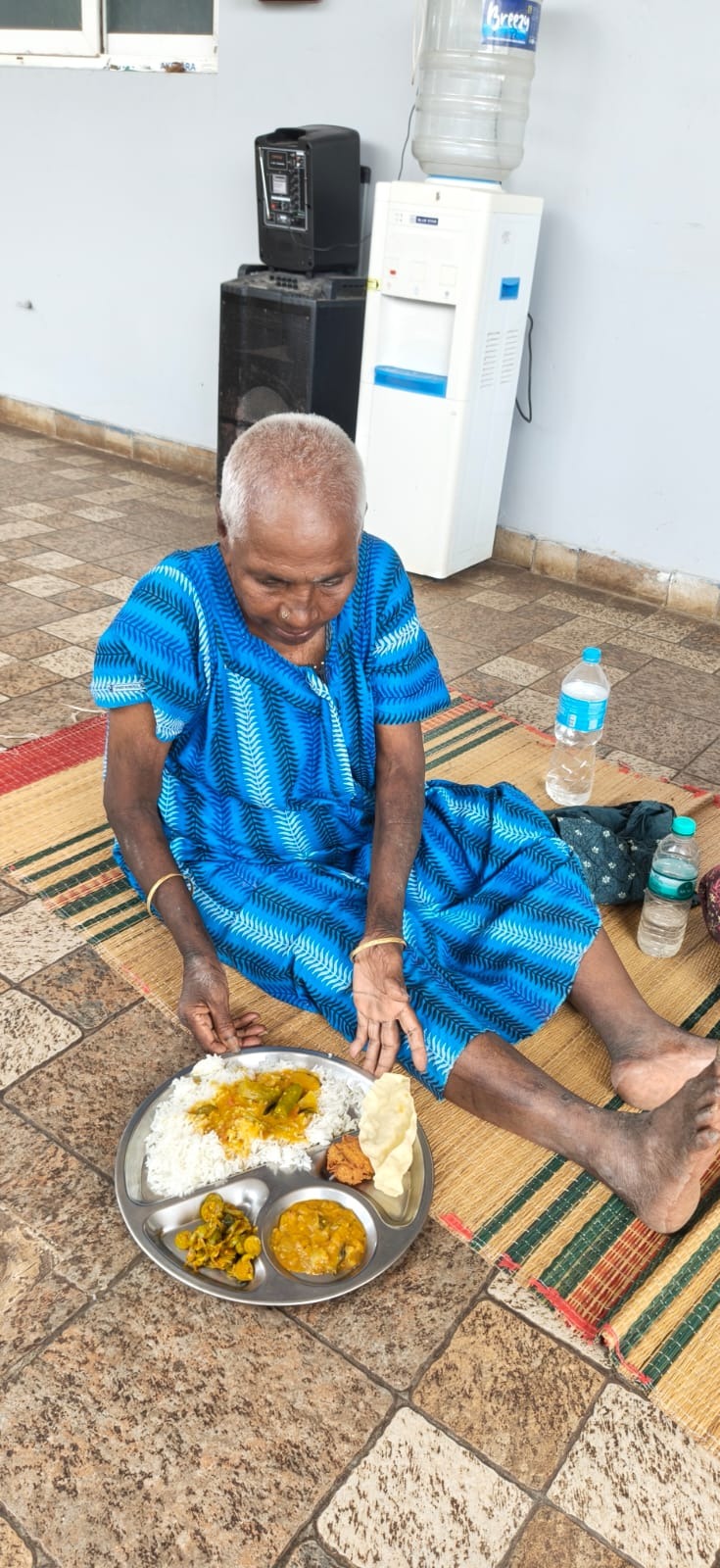 Gangammal seated on a mat at the shelter with a plate of rice, curry and papad