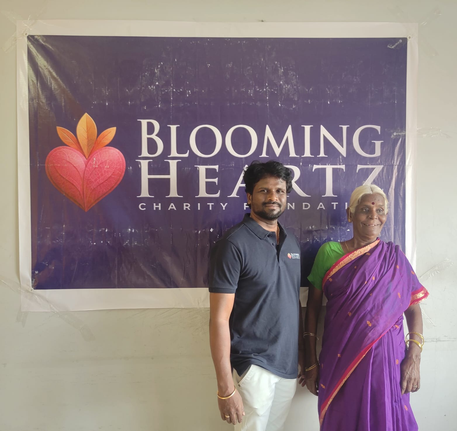 A recovered and smiling Buchamma standing with a volunteer in front of the foundation banner